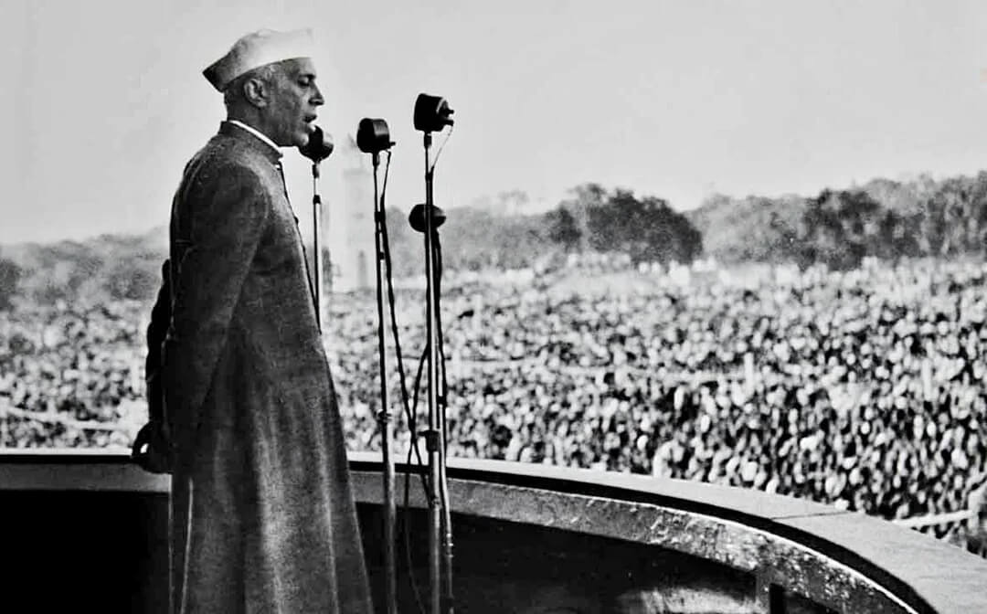 Jawaharlal Nehru standing at a podium before a vast crowd, with microphones lined up in front.