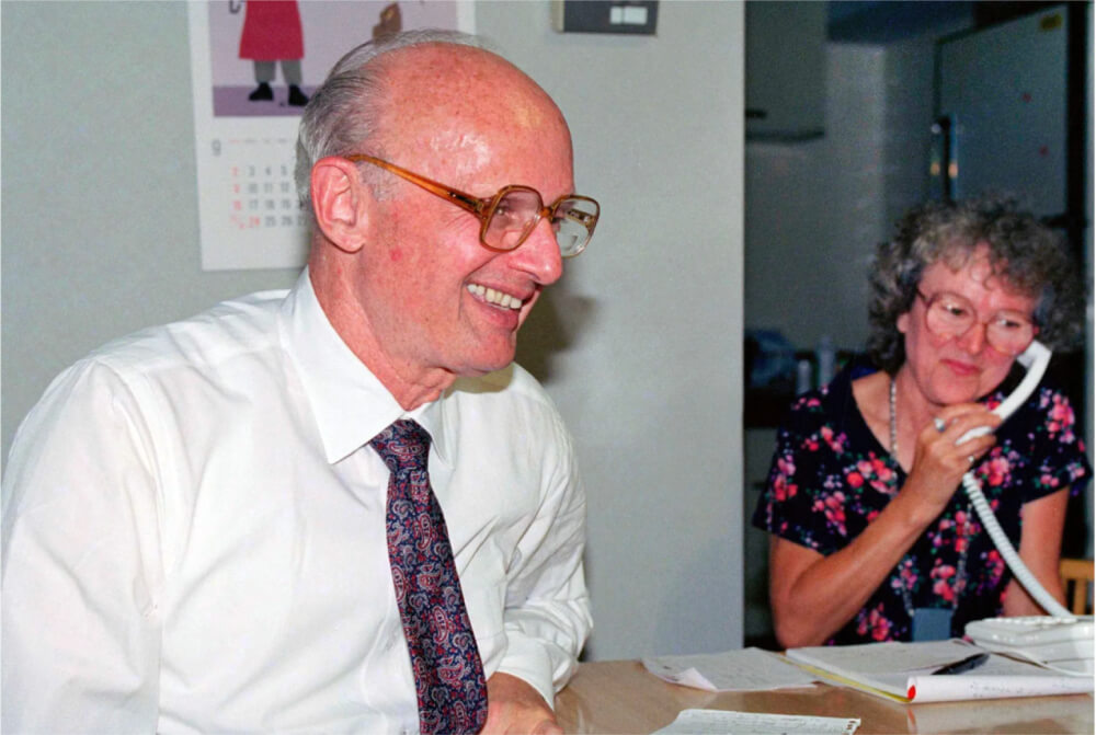 Harry Markowitz seated at his desk, smiling, with a colleague on a phone call nearby